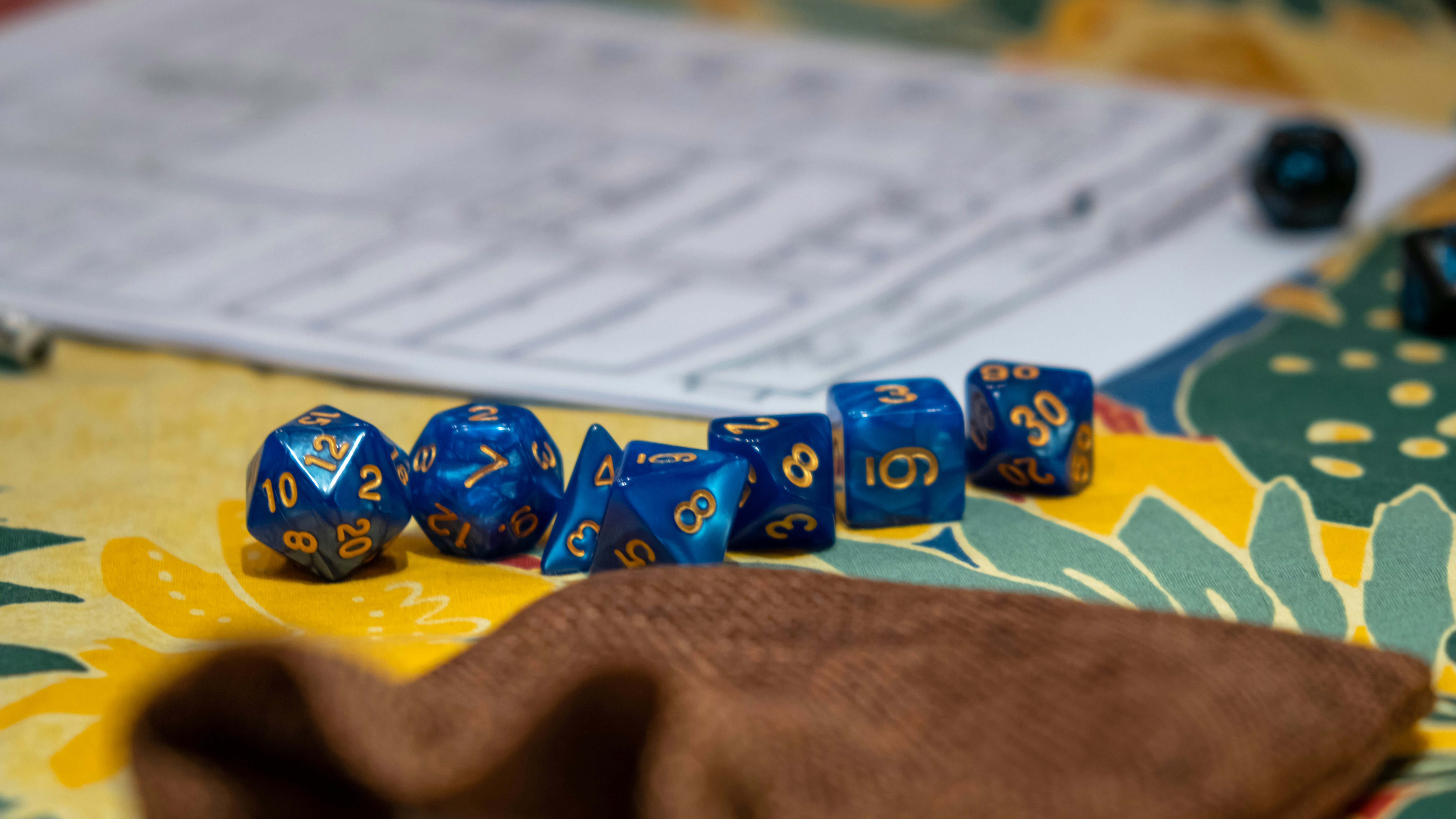 A set of metallic blue polyhedral dice with gold lettering are lined up on a table. A dice bag is in the foreground and a character sheet in the near distance.
